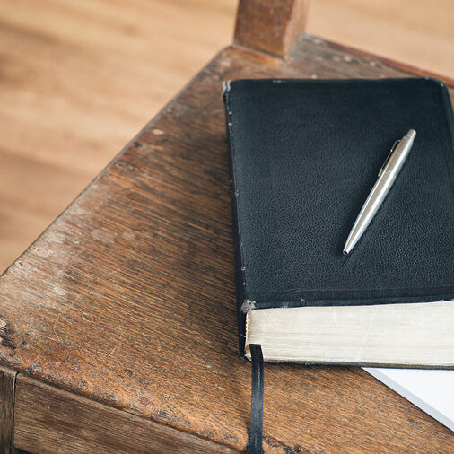 A black journal with a pen rests over an open paper notebook on an old wooden chair.